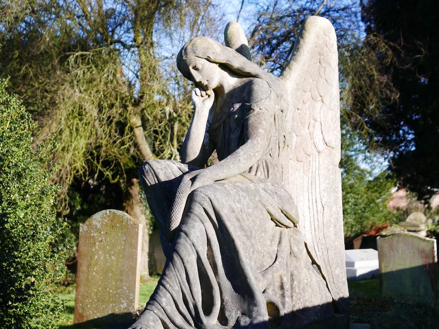 A weathered stone statue of an angel seated in a thoughtful pose within a graveyard. The angel rests its chin on one hand while holding a scroll across its lap, wings rising gracefully behind. Sunlight highlights the detailed carving, with gravestones and leafless trees visible in the background.