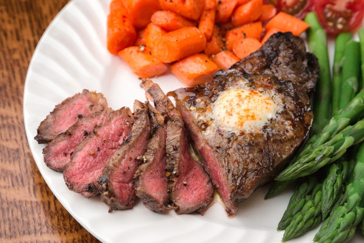 Ribeye steak with butter and asparagus and carrots on the side on a white plate.