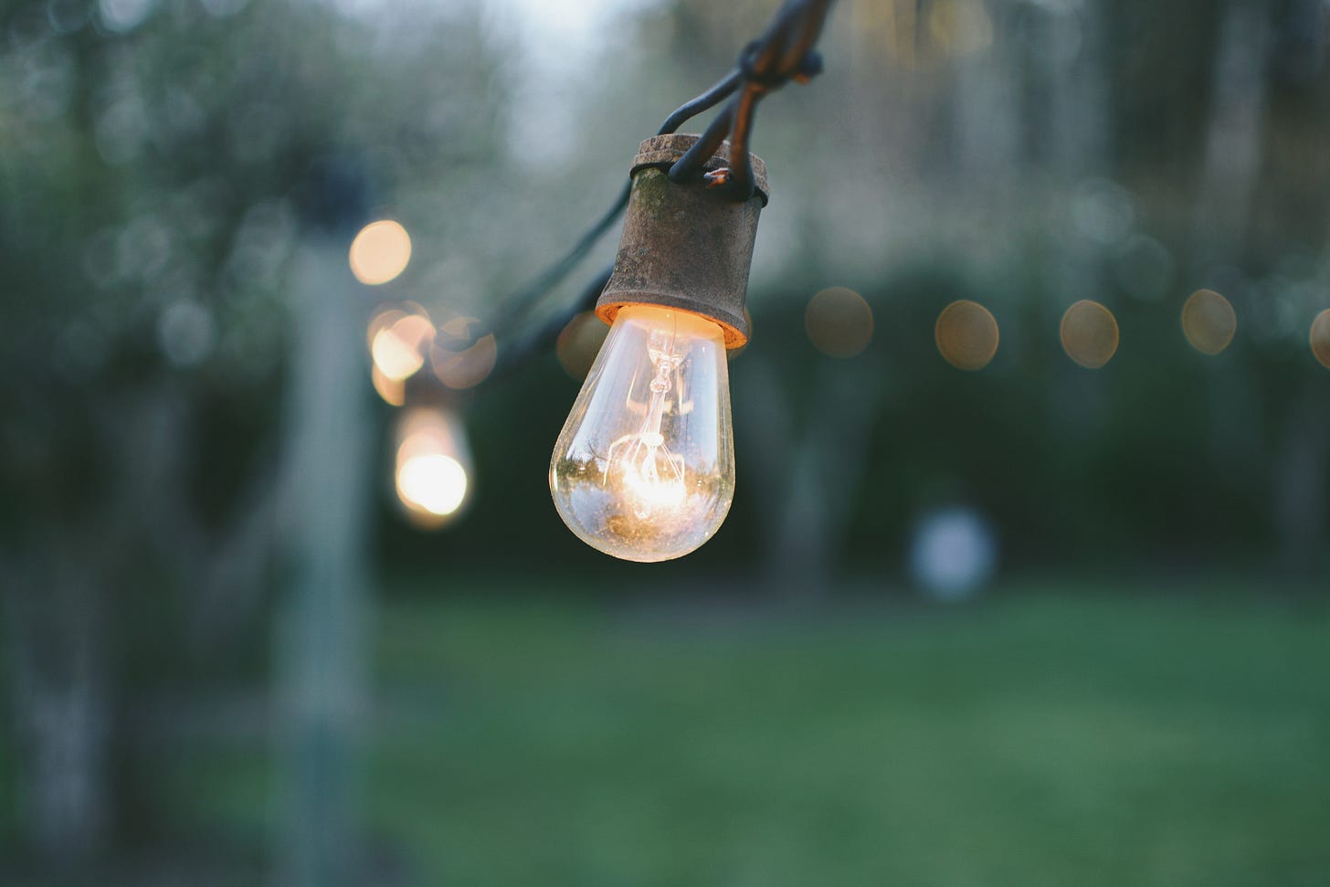 Primo piano di una lampadina accesa appesa a un filo di lampadine sopra un prato sfocato sullo sfondo.