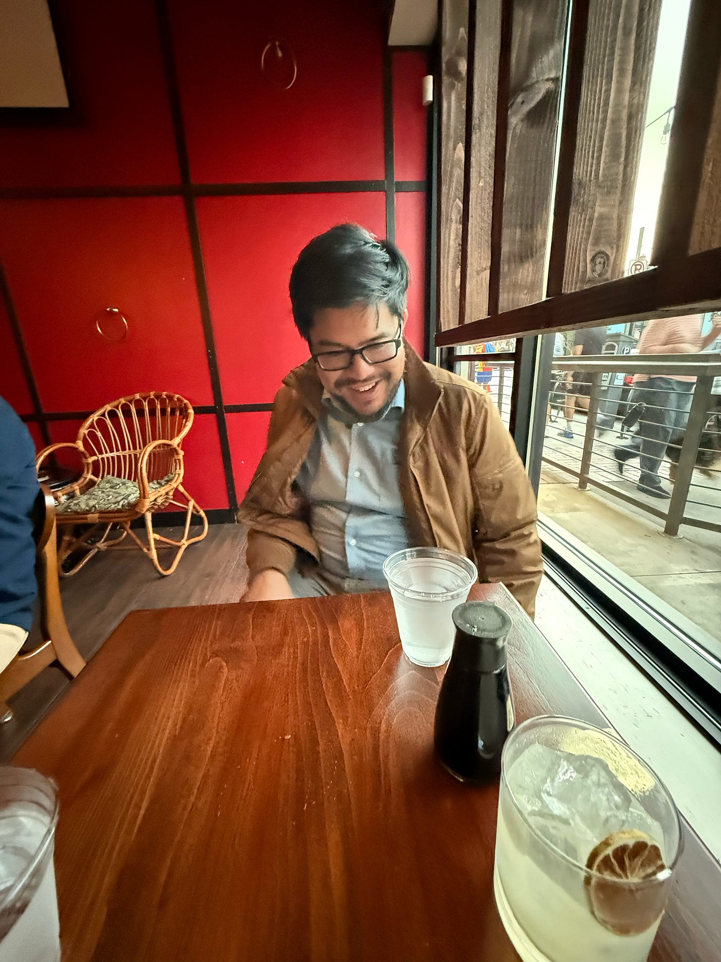 William (with dark hair and glasses, wearing a jacket and button-up shirt) sits opposite the interviewer at a table in a restaurant.