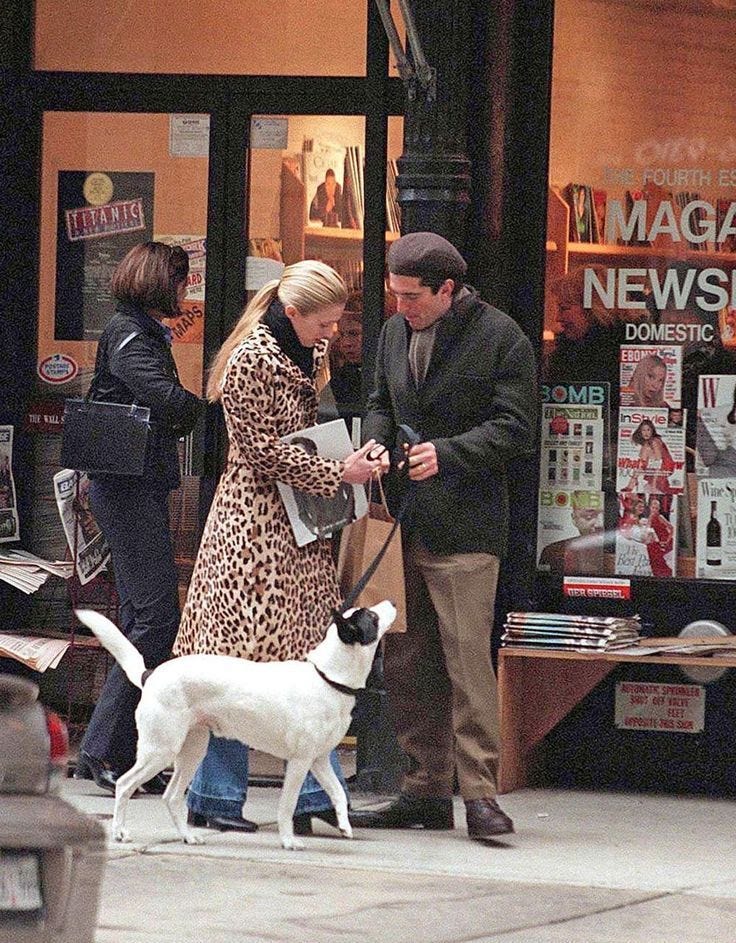 This may contain: a man and woman standing on the sidewalk with a dog in front of a store