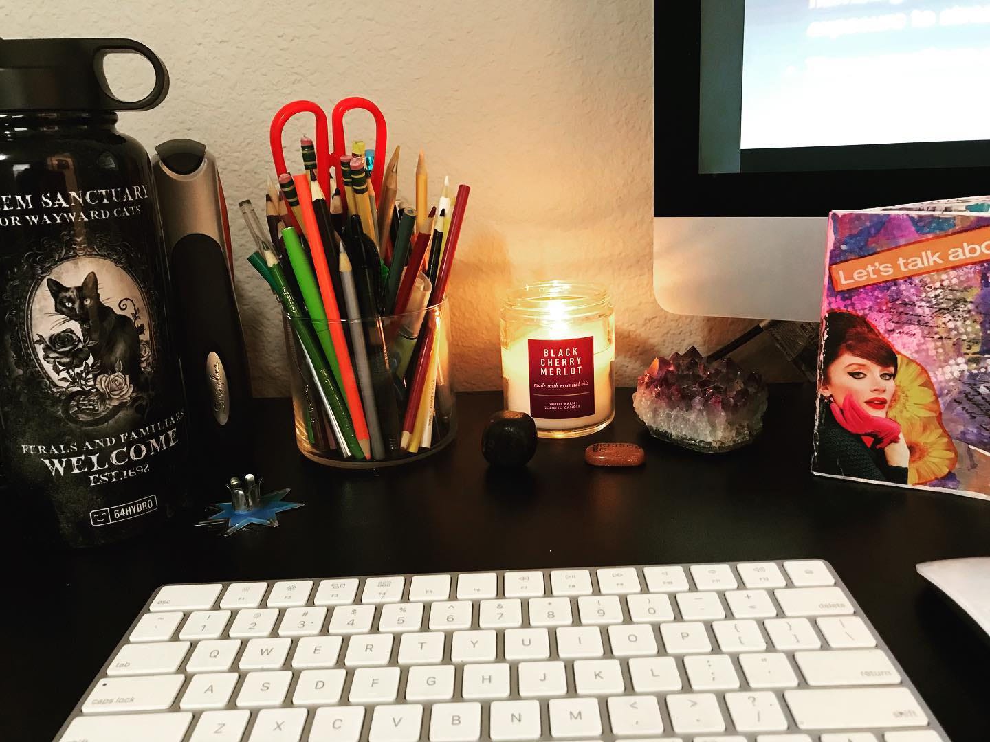 Keyboard and computer screen, surrounded by jar of pens, a water bottle, a burning candle, various gemstones, and small art zine