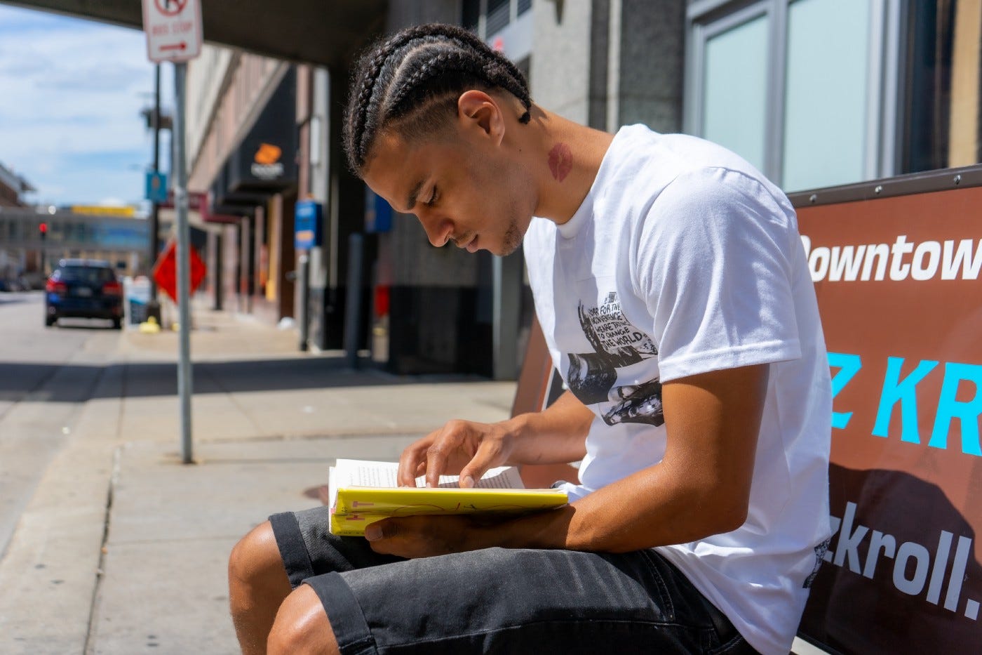 A casually dressed young man reads a book on a city bench. He is engrossed in the reading. Urban landscape of signs, buildings, and cars are in the background. A casually dressed young man reads a book on a city bench. He is engrossed in the reading. Urban landscape of signs, buildings, and cars are in the background.