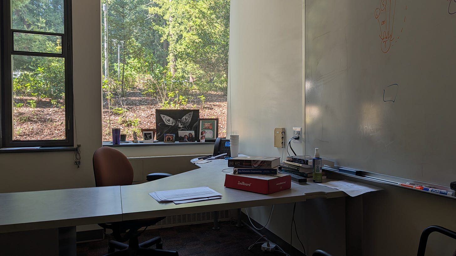 A faculty office with a large window looking onto redwoods, a whiteboard, books, family photographs, and an FPGA development board on the desk.