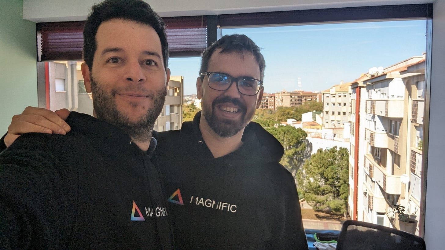 Two people wearing black Magnific hoodies posing for a selfie in front of a large office window.