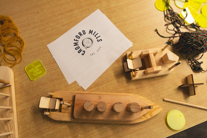 Overhead view of two handmade wooden boats on a wooden table next to a card that says Cromford Mill, and balls of strings
