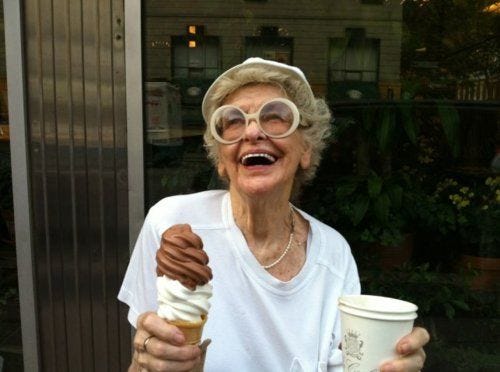 This may contain: an older woman holding up a cup of ice cream and a chocolate covered donut