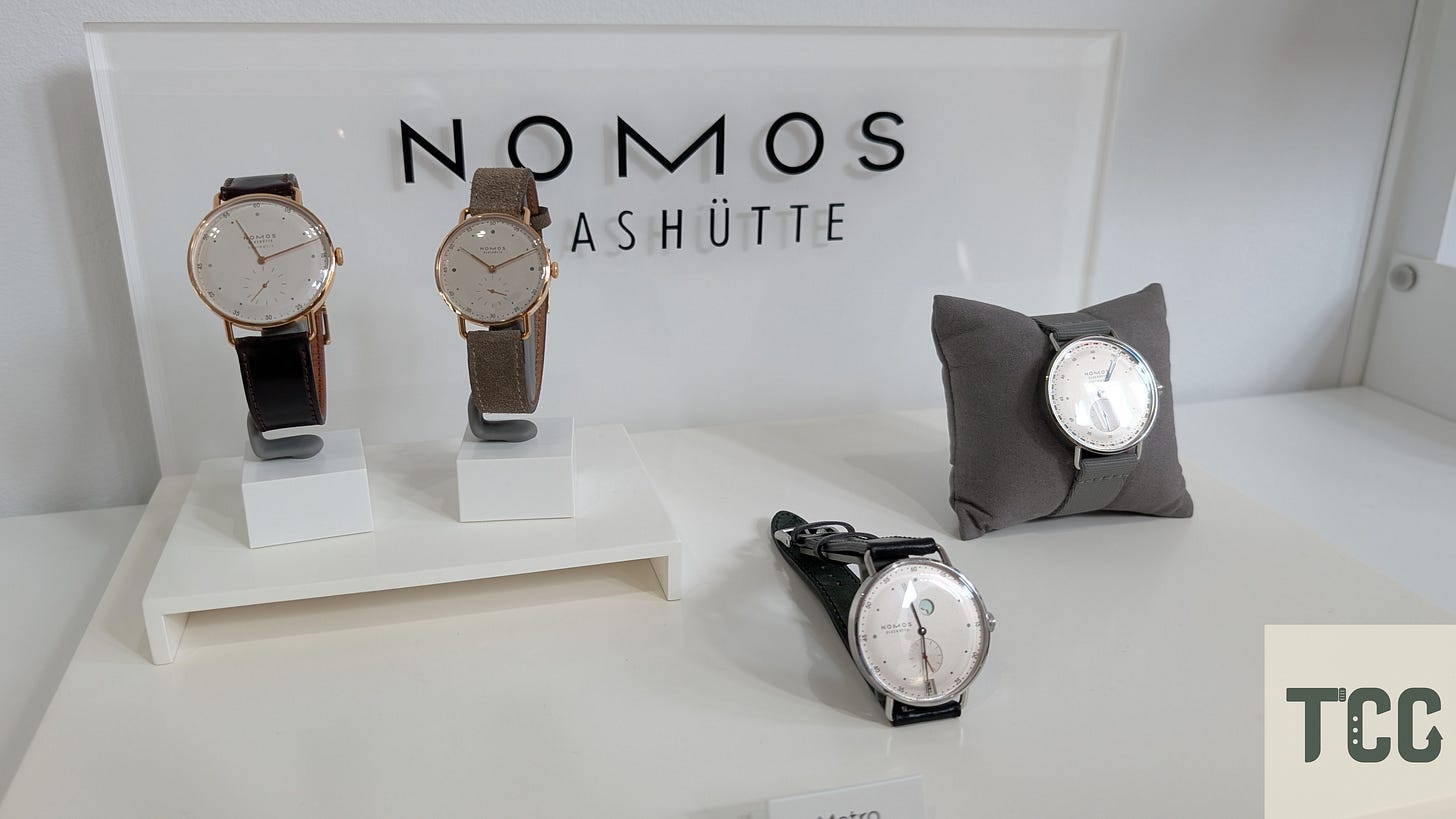 Four NOMOS watches on display including Metro and Orion models, with sleek dials, leather straps, and minimalistic details, placed on white and gray stands in a boutique setting.