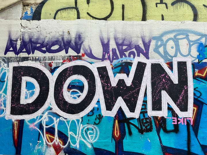 Black, white, and pink "DOWN" on a graffiti wall, over 100 handwritten "aesthetic" stickers on a rolling chair, "Like" painted in blue, yellow, and white facing up and down, and "motherquake" painted in black on a yellow square.