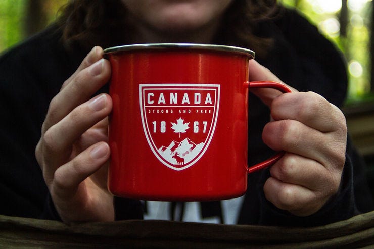 Person holding a red mug with "CANADA, STRONG AND FREE, 1867" text. Dark clothing, blurred green forest background. Person holding a red mug with "CANADA, STRONG AND FREE, 1867" text. Dark clothing, blurred green forest background.