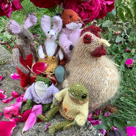 Knitted toys under a red rose bush surrounded by fallen petals
