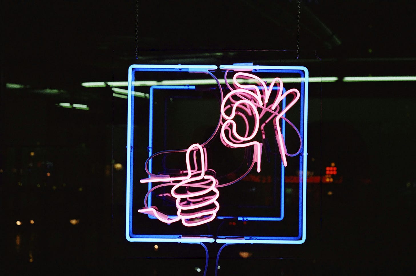 A square of green and blue neon outline with pink neon hands: one with thumb up and the other has thumb and forefinger held in 'ok' gesture.