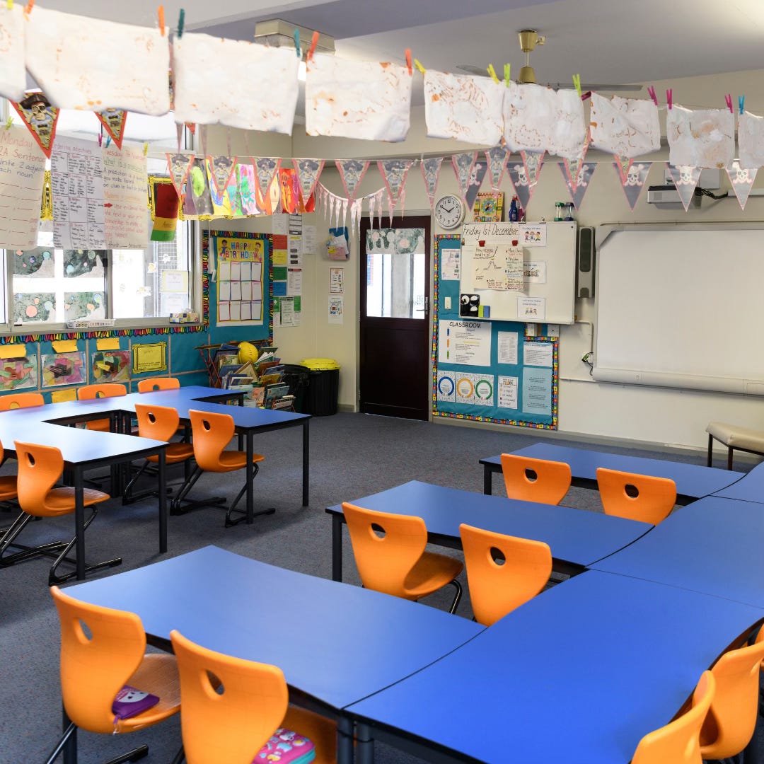 Interior of a primary classroom. There are blue tables with orange chairs and multi-coloured display boards