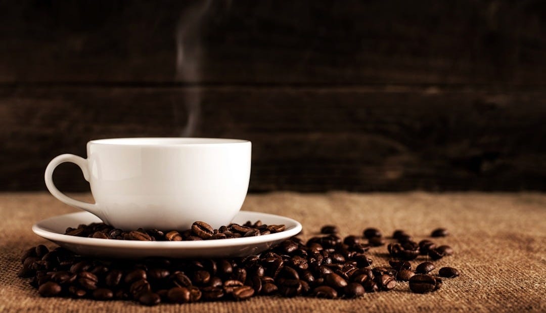 white ceramic mug and saucer with coffee beans on brown textile