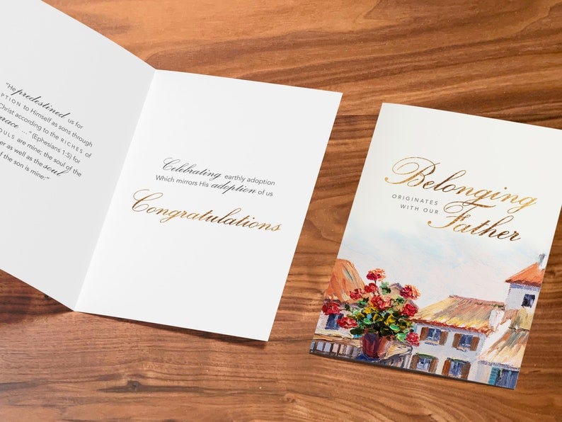 May include: A folded card with a gold inscription that reads 'Celebrating earthly adoption which mirrors His adoption of us Congratulations.' The card has a watercolor painting of a cityscape with red flowers in a pot on a balcony. The inscription on the front of the card reads 'Belonging originates with our Father.'