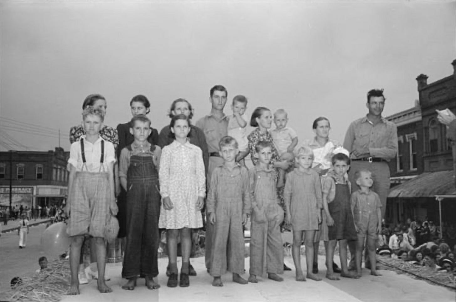 A black and white photo of a family of 18 on a stage, in a small town's Main Street during a celebration. At least nine of the kids are barefoot. A black and white photo of a family of 18 on a stage, in a small town's Main Street during a celebration. At least nine of the kids are barefoot.
