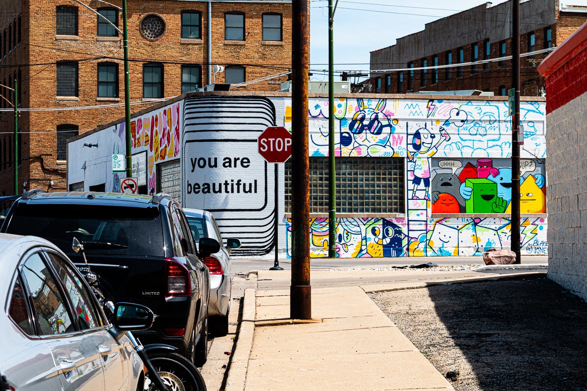Street view showing a colorful mural with playful characters and a large “you are beautiful” painted message on a wall, next to parked cars and a stop sign. Street view showing a colorful mural with playful characters and a large “you are beautiful” painted message on a wall, next to parked cars and a stop sign.