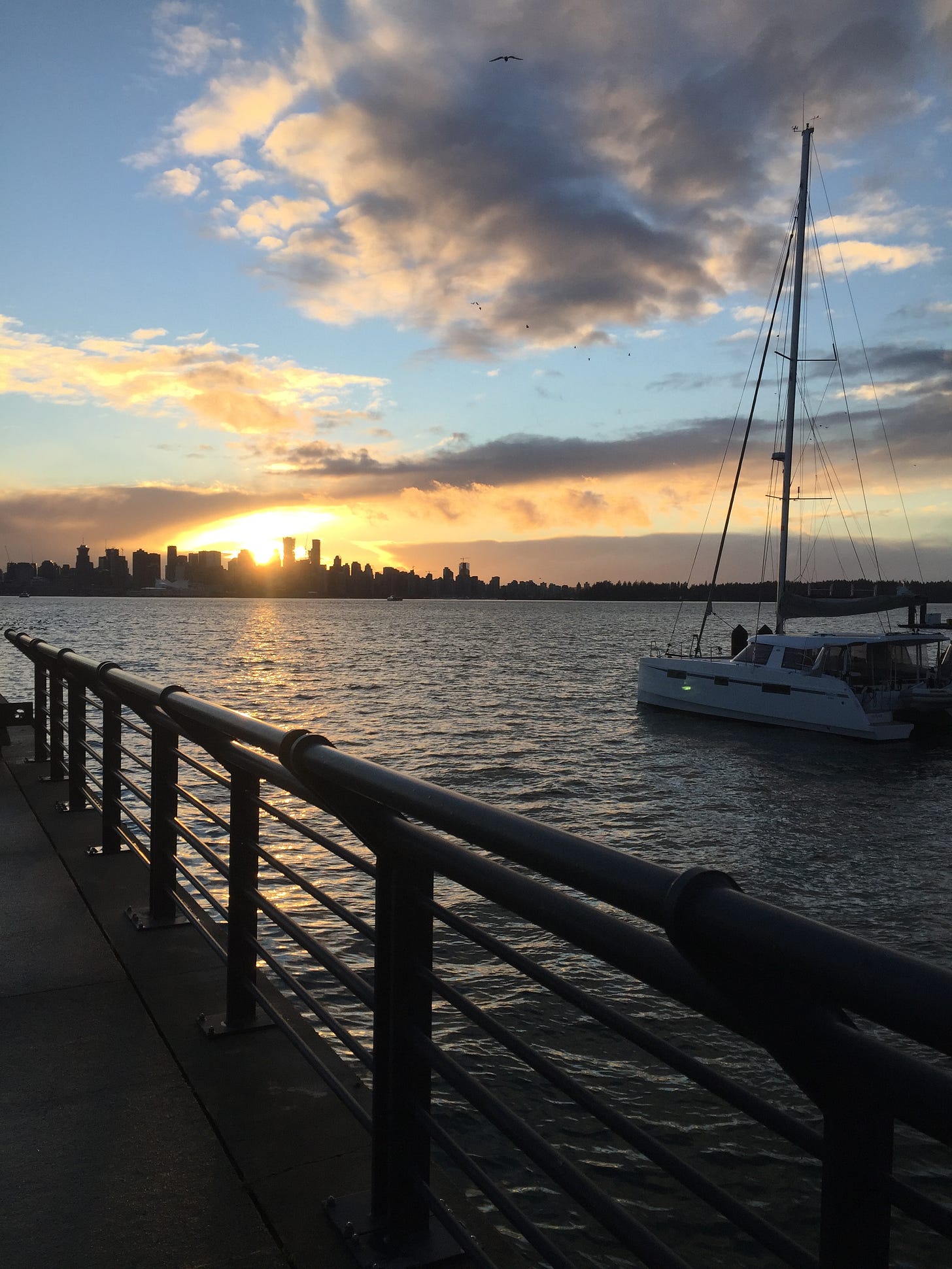 A view of Vancouver from the Lonsdale Pier. The sun is setting over the city and scattered clouds darken in a blue sky. A sailboat is in the harbour next to the pier.