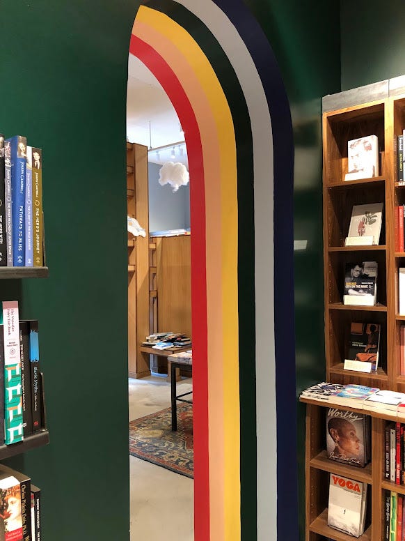 A slightly tilted view of a rounded archway. The walls are a dark green, and the interior of the archway is a rainbow. In front of the archway are some wooden bookshelves, and beyond the archway, more bookshelves, with clouds hanging from the ceiling. 