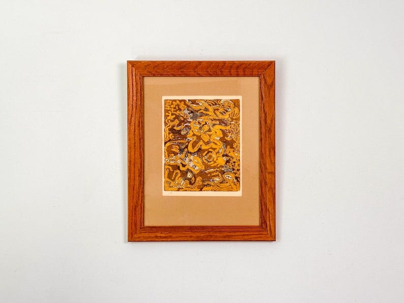 May include: A framed artwork with a brown wooden frame. The artwork features a detailed, abstract design in shades of gold and brown. The artwork is set against a tan mat.