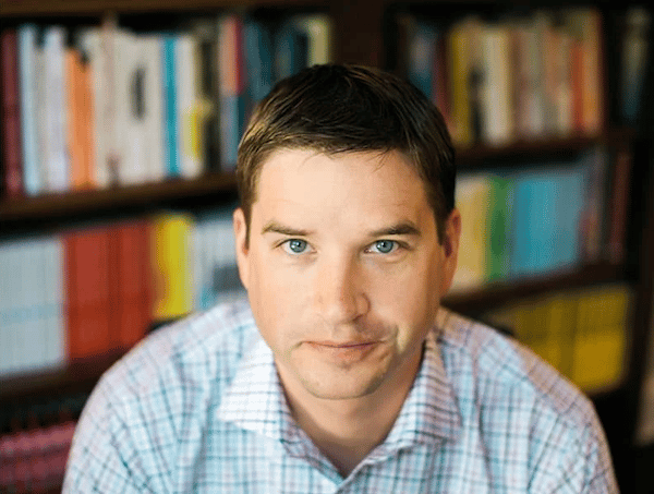 A man with short, dark hair and wearing a checked shirt, looking directly at the camera with a neutral expression, in front of a bookshelf filled with various books.