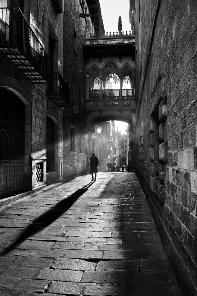 A grayscale photograph with strong contrasts. There are darkened walls, the sun shining down, and a man at the focal point, his shadow casting a large contrast with everything else. A grayscale photograph with strong contrasts. There are darkened walls, the sun shining down, and a man at the focal point, his shadow casting a large contrast with everything else.