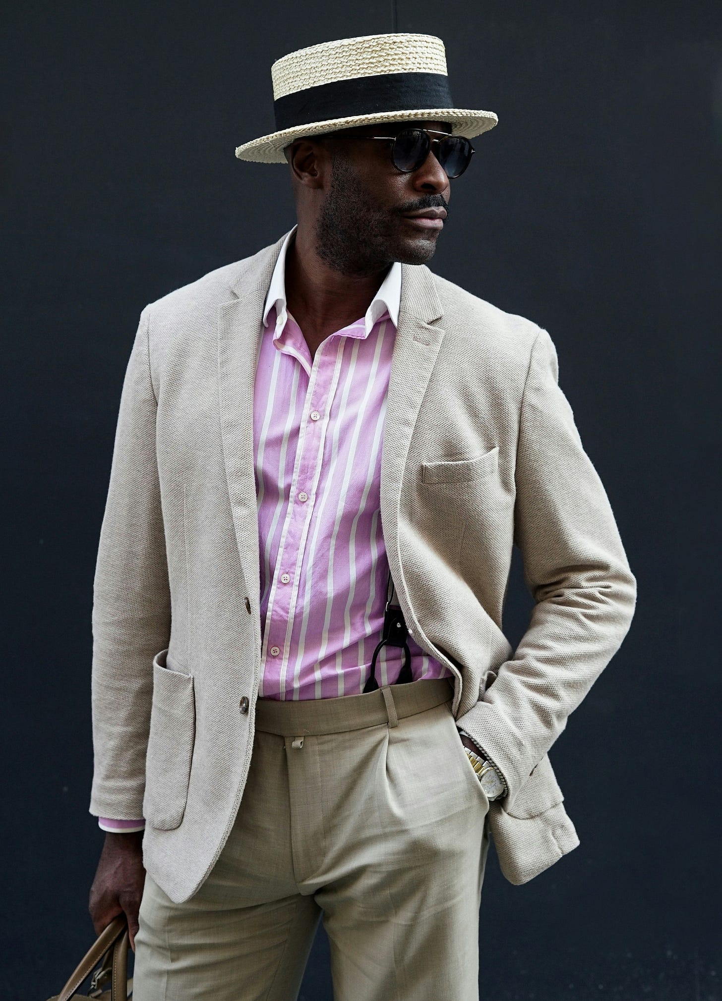 Man wearing classic Black Dandy fashion with straw boater hat, light beige linen suit, pink striped shirt and sunglasses against dark background