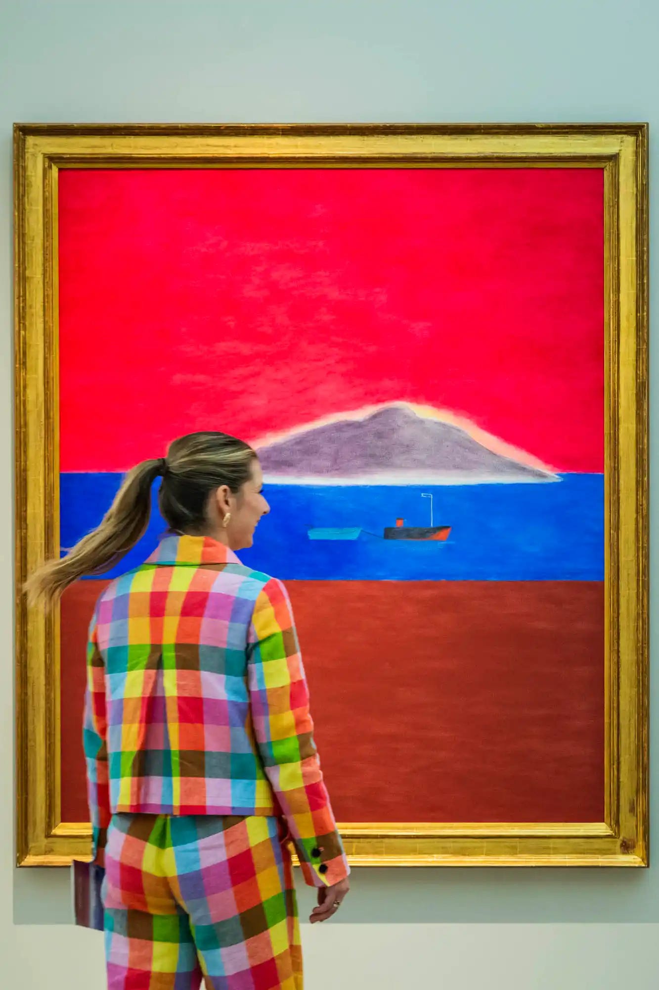 A visitor in a colorful checkered suit admires a vibrant seascape painting featuring a red sky and calm blue sea at the British Art Fair 2025.