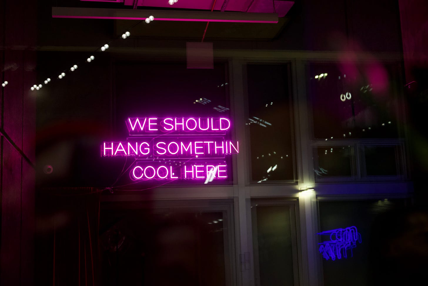 A magenta neon sign reading “WE SHOULD HANG SOMETHIN COOL HERE” mounted against windowed walls at night, its glow reflecting across glass panes while soft motion blur and foreground haze create layered depth and a saturated, cinematic atmosphere.