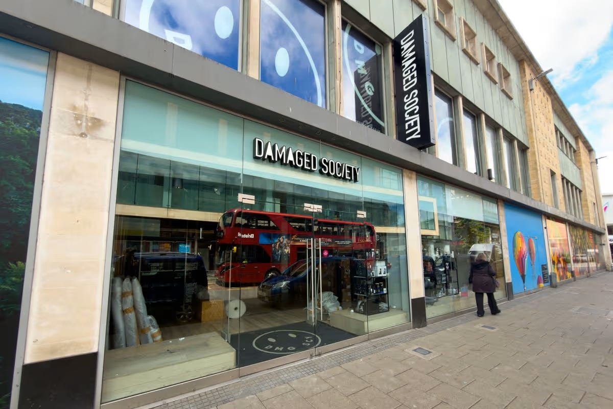 Damaged Society clothing store in Broadmead, Bristol city centre — black "Damaged Society" signage and smiley logo above a glass shopfront, a red Bristol bus reflected in the windows. The Almighty Gob, www.thealmightygob.com
