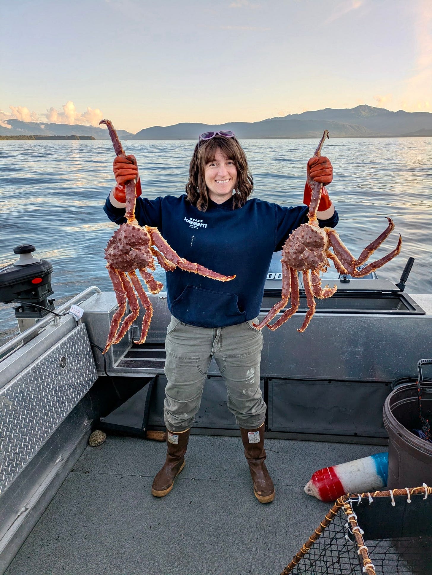 Michelle standing on a boat and holding up two king crabs with the SE Alaska water and landscape behind her.