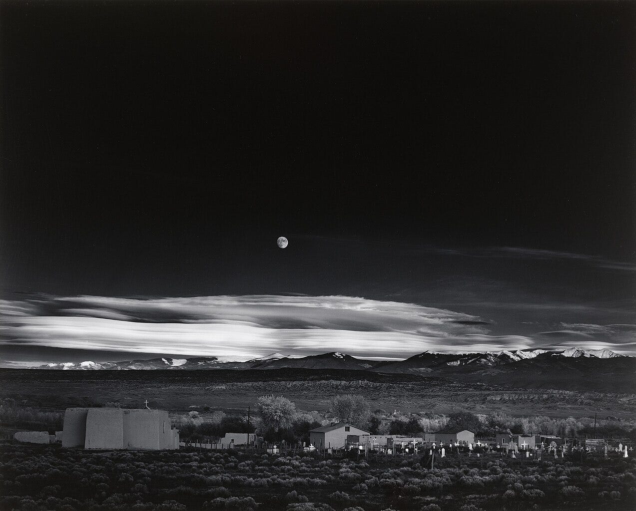 Ansel Adams' iconic black-and-white photograph 'Moonrise, Hernandez, New Mexico,' taken on November 1, 1941, showing a dramatic landscape with the moon rising over a small village, snow-capped mountains in the background, and clouds illuminated by the setting sun.