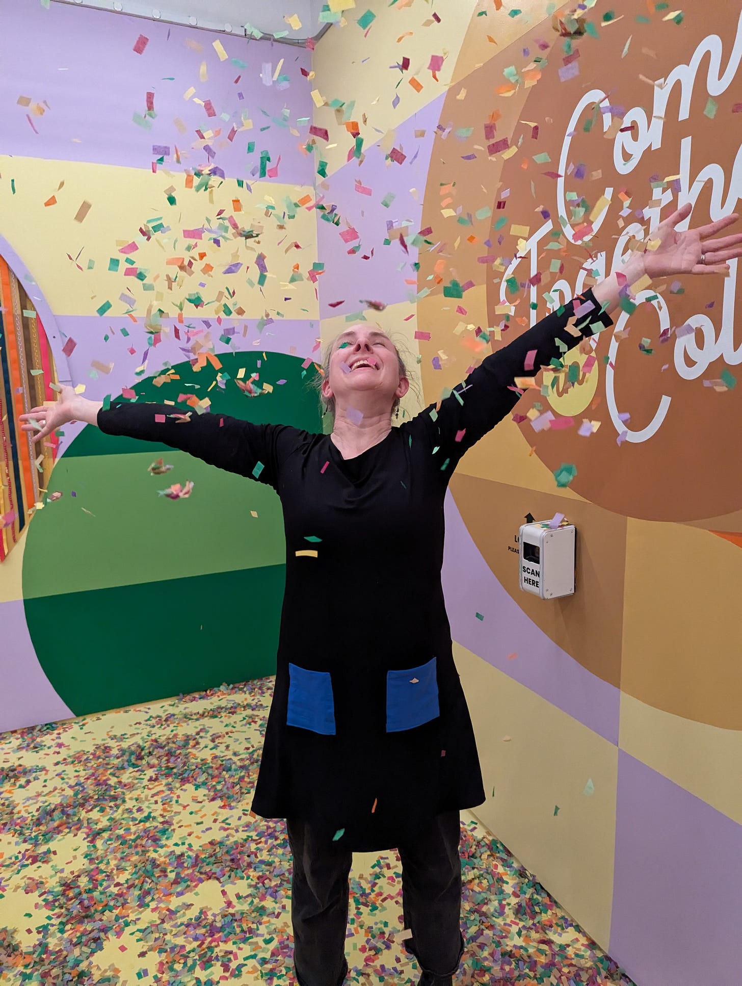 A middle-aged white woman at The Color Factory, throwing confetti in the air and smiling up at it. A middle-aged white woman at The Color Factory, throwing confetti in the air and smiling up at it.
