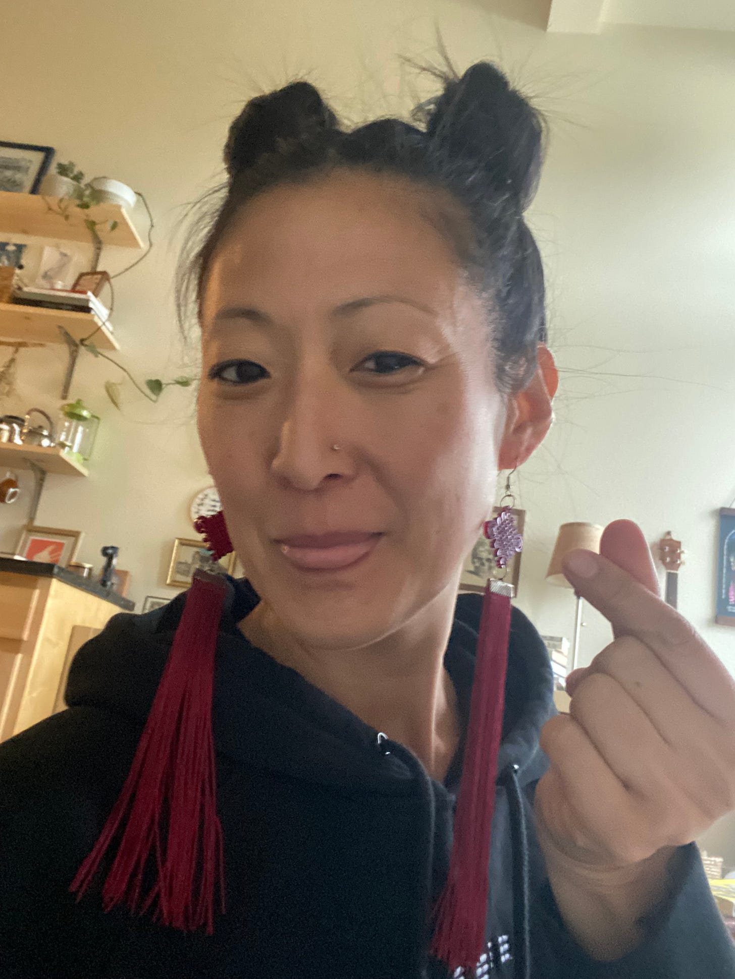 A woman with space buns and long flowy red earrings with a black sweatshirt on with her tongue playfully sticking out while she's giving a heart-finger. The background is a beige wall with some wooden shelves to the left. A woman with space buns and long flowy red earrings with a black sweatshirt on with her tongue playfully sticking out while she's giving a heart-finger. The background is a beige wall with some wooden shelves to the left.