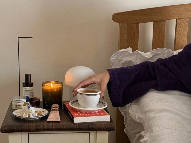 This may contain: a person sitting on a bed next to a night stand with a cup of coffee This may contain: a person sitting on a bed next to a night stand with a cup of coffee