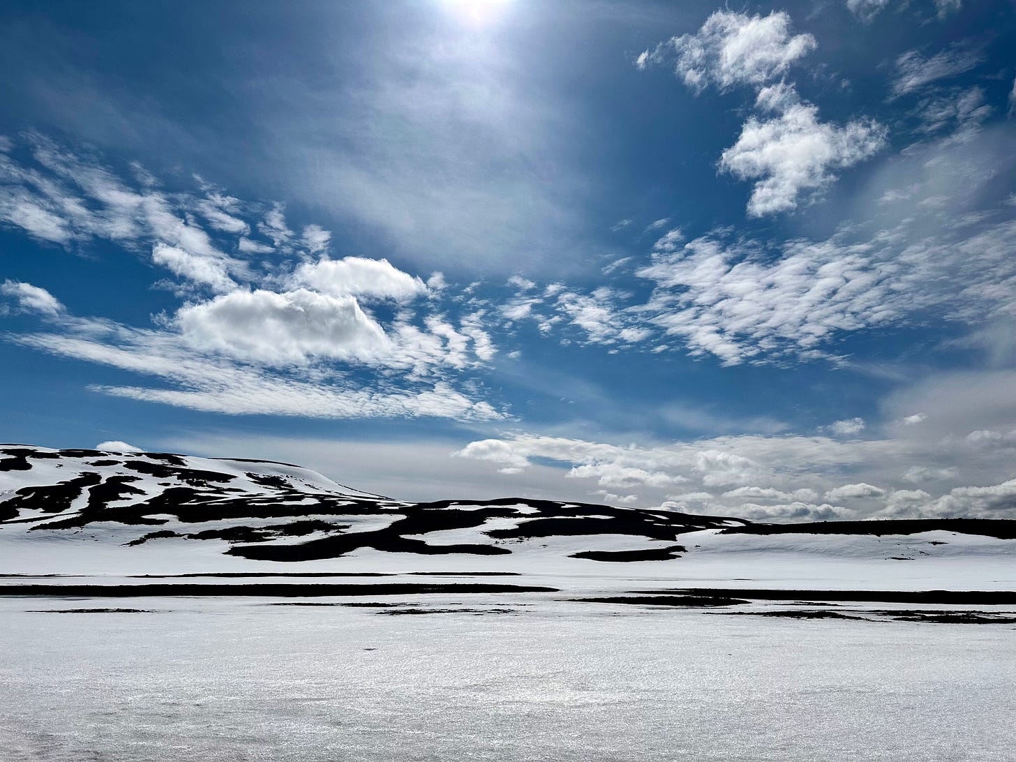 Snow covered ground with snowy mountains in the distance under a blue sky with puffy, white clouds.  Iceland, May 2024