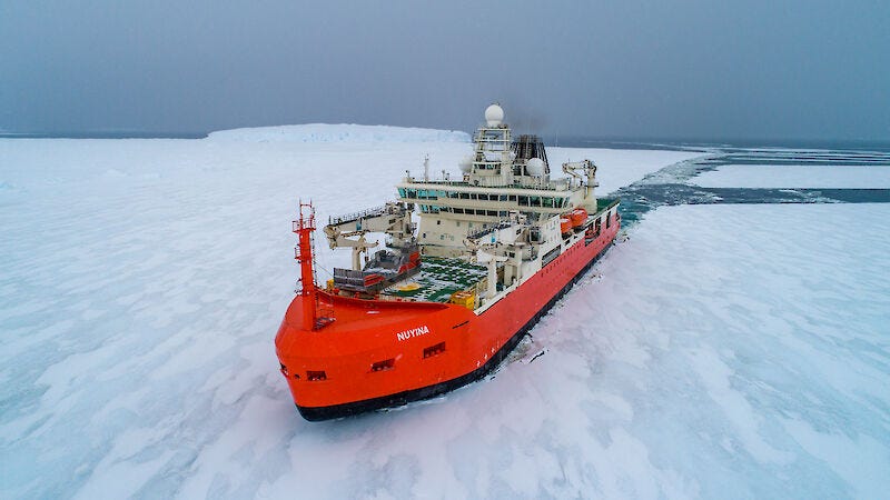 An aerial drone photo of an orange and white ship breaking through sea ice. There is a long channel of water behind the ship where it has broken a path through the white expanse of the ice. An aerial drone photo of an orange and white ship breaking through sea ice. There is a long channel of water behind the ship where it has broken a path through the white expanse of the ice.