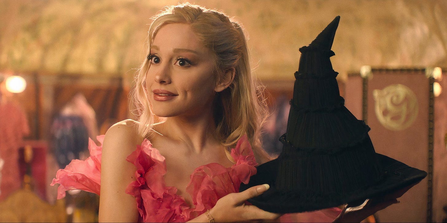 Ariana Grande in Wicked as Galinda, holding up a black pointy hat to offer to Elphaba.