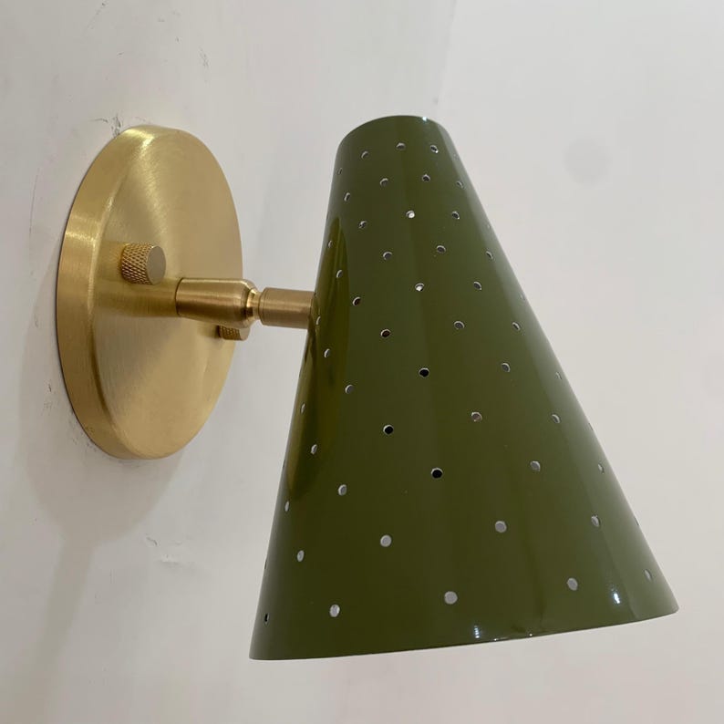 May include: Olive green cone-shaped wall sconce with a perforated design. The sconce features a brass-colored circular base and adjustable arm. The fixture is mounted on a white wall, offering a modern lighting solution. May include: Olive green cone-shaped wall sconce with a perforated design. The sconce features a brass-colored circular base and adjustable arm. The fixture is mounted on a white wall, offering a modern lighting solution.