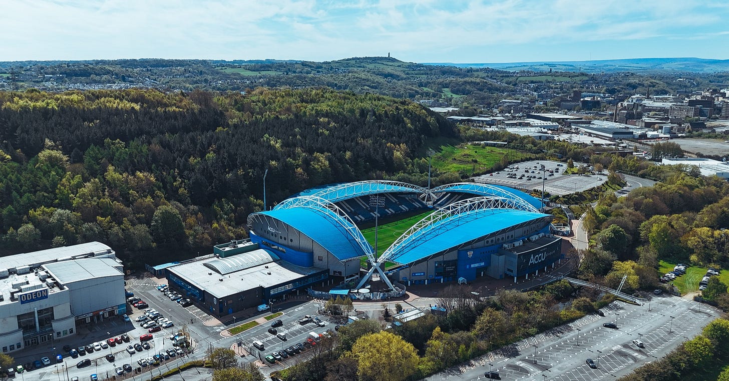 A photograph of Huddersfield Town’s Accu Stadium on a bright, sunny day. The town centre (right) and Victoria Tower (left) can be seen in the background.