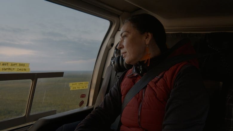An Indigenous woman with brown hair looks out the window of a helicopter. 