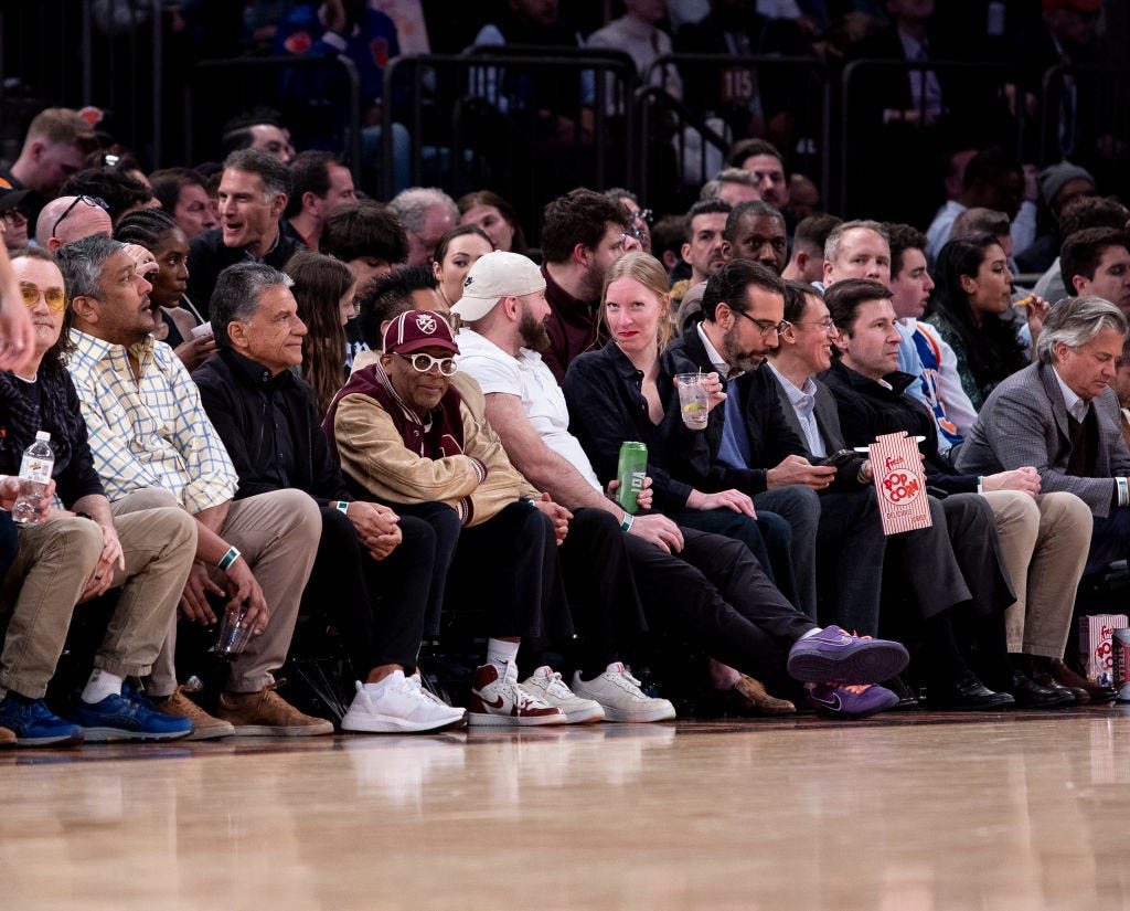 Director Spike Lee sits courtside at an arena basketball game, wearing round white-rimmed glasses, a maroon baseball cap, and a maroon varsity jacket with tan sleeves alongside a row of spectators watching the court.