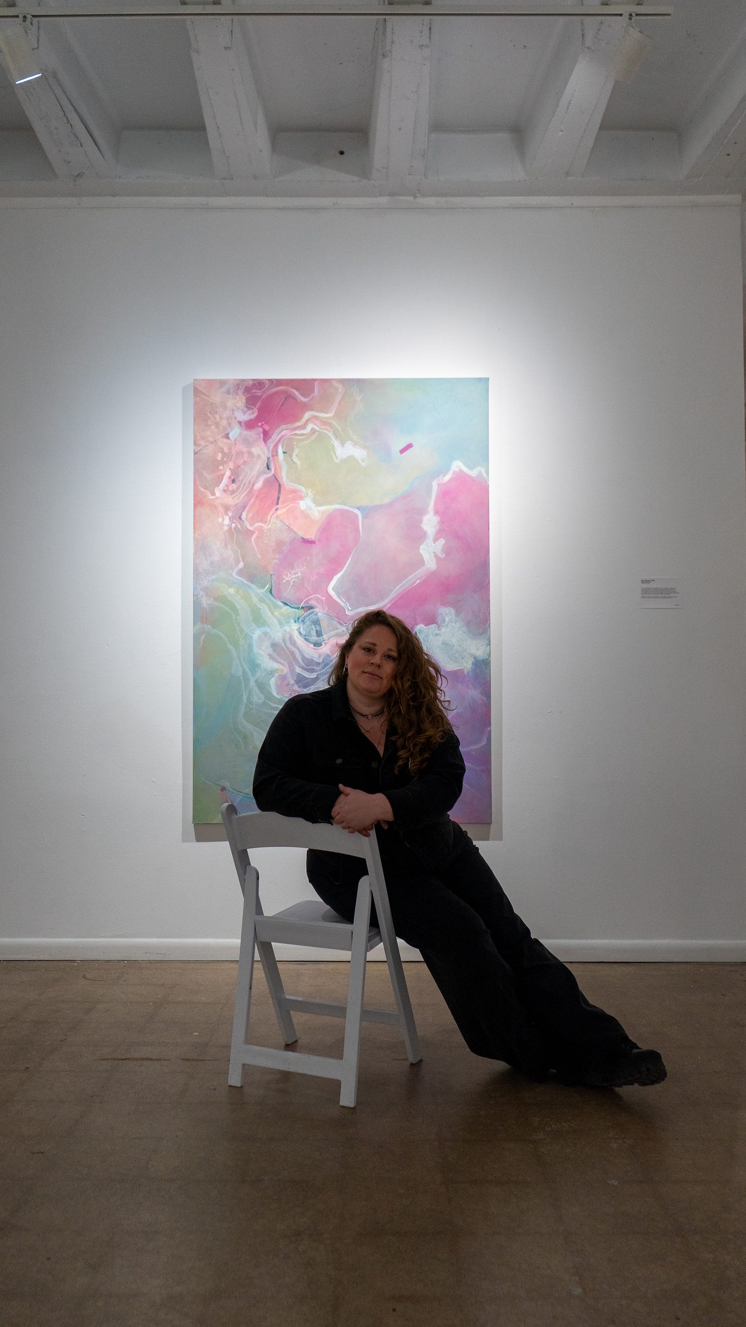 This portrait-oriented photo shows Briget sitting in a white folding chair in front of one of her Aha Moment paintings. This painting is primarily pastel with soft swirling pinks, blues, and greens with white ripples. Briget wears a dark jumpsuit and has long curly red hair. She smiles softly at the camera. This portrait-oriented photo shows Briget sitting in a white folding chair in front of one of her Aha Moment paintings. This painting is primarily pastel with soft swirling pinks, blues, and greens with white ripples. Briget wears a dark jumpsuit and has long curly red hair. She smiles softly at the camera.
