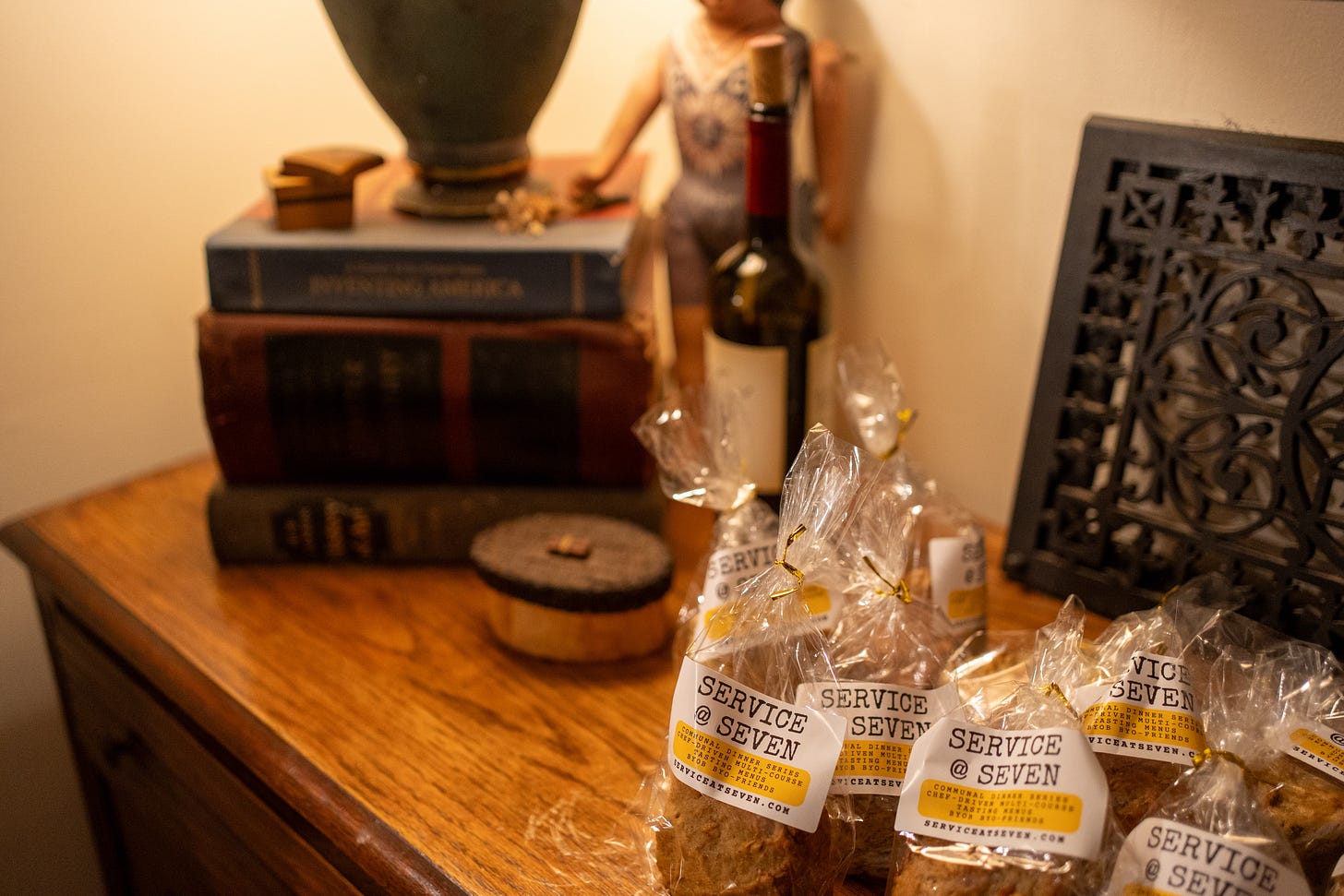 Clear plastic bags tied with string holding small loaves of bread, labeled with yellow Service @ Seven stickers, resting on a wooden surface next to stacked vintage books and a wine bottle.
