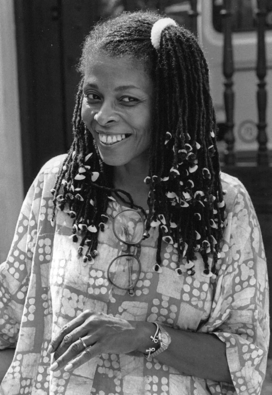 B&W image of Assata Shakur. She is smiling and looking at something to the right of the camera. Her locs are half up half down in a side ponytail, with beads on the ends. She wears an African print tunic and simple jewelry. Her glasses hang from her collar.
