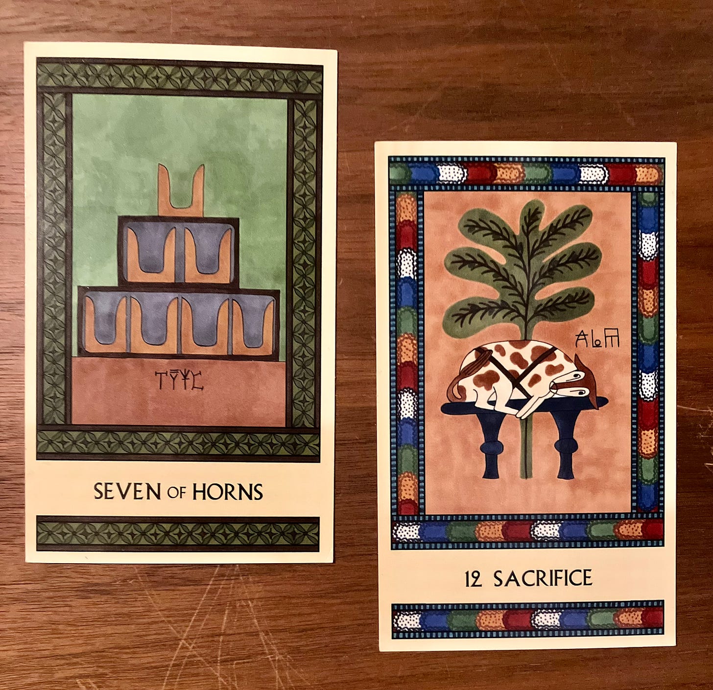 Two Minoan Tarot cards side by side on a dark brown wood surface. The Seven of Horns is painted in shades of green and brown. It shows seven pairs of Minoan sacred horns sitting on a plain, unadorned shrine. Sacrifice has a bright multicolored border. It shows a spotted bull trussed on a table in preparation for sacrifice, with an olive tree standing behind it.