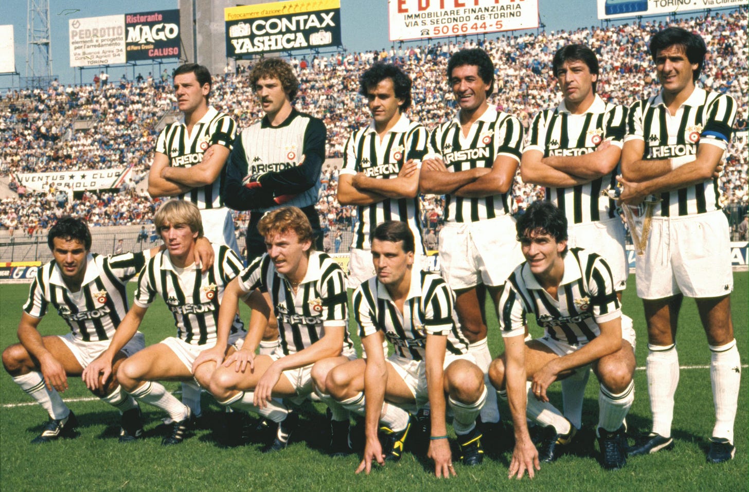 Juventus starting lineup in the 1980s wearing Ariston jerseys, with Michel Platini and teammates posing before a match at a packed stadium.