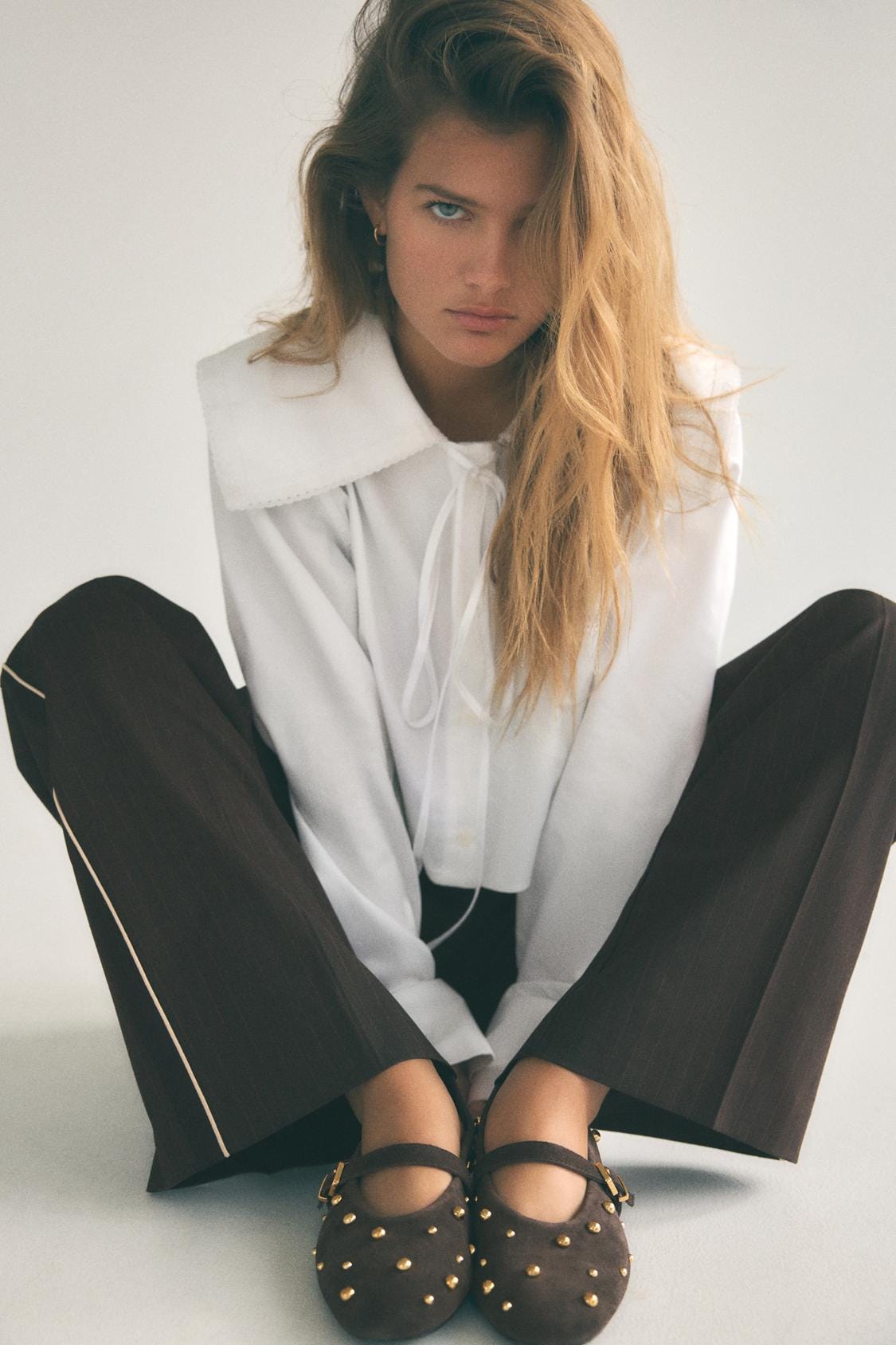 White blouse with a large collar, paired with brown trousers and brown studded shoes.