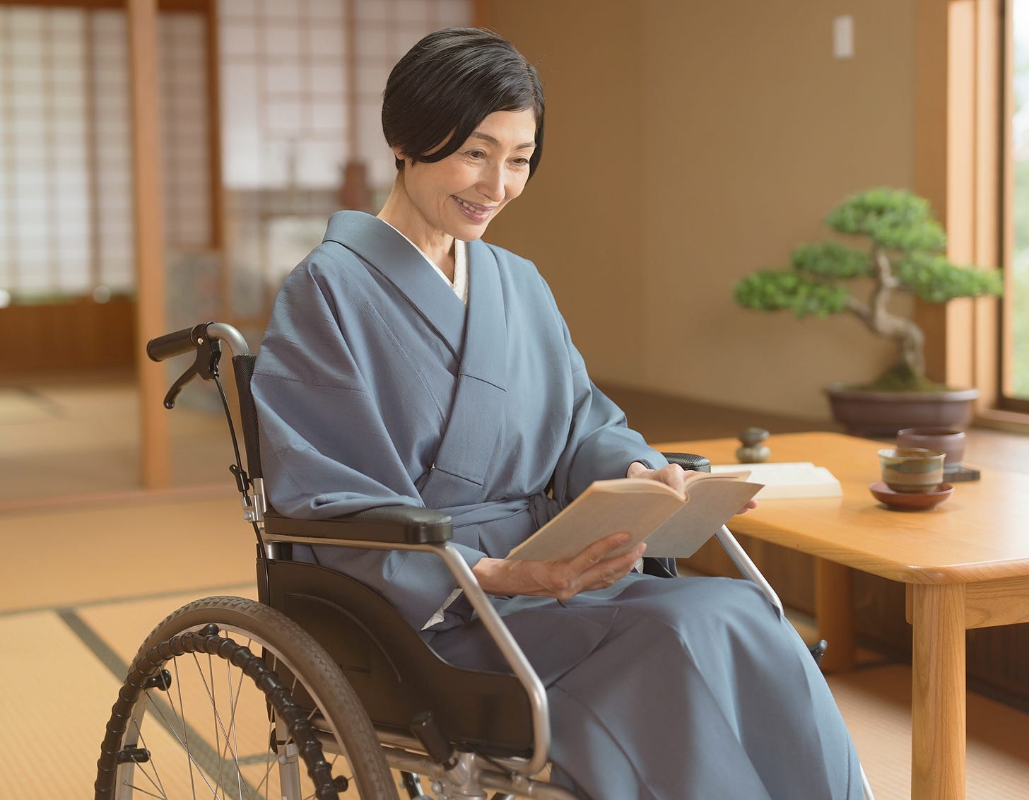A middle-aged woman wearing an elegant soft blue kimono sits comfortably in a wheelchair, smiling gently as she reads a book in a peaceful Japanese-style room. Warm natural light filters through shoji screens in the background, casting a calm atmosphere over the tatami-floored space. Beside her, a low wooden table holds ceramic tea bowls and a small, carefully pruned bonsai tree, adding to the quiet sense of tranquility in the scene.