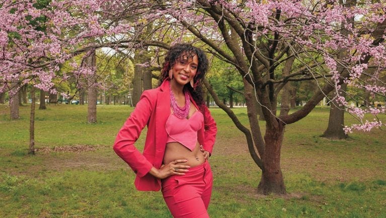 Polyamory influencer Millie Boella poses in a park next to a cherry blossom tree, wearing a hot pink suit